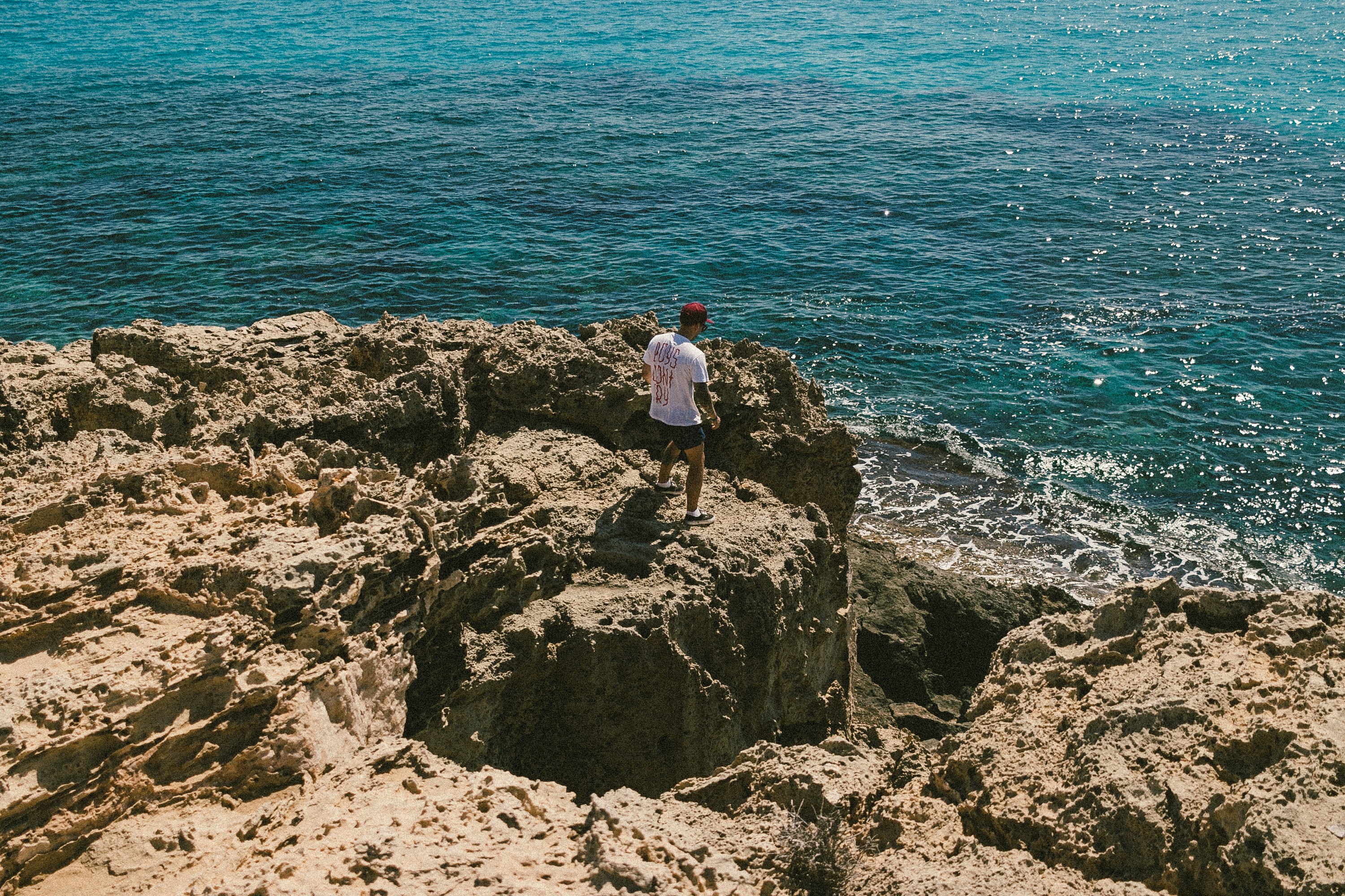 man in white t-shirt standing on rock formation near body of water during daytime